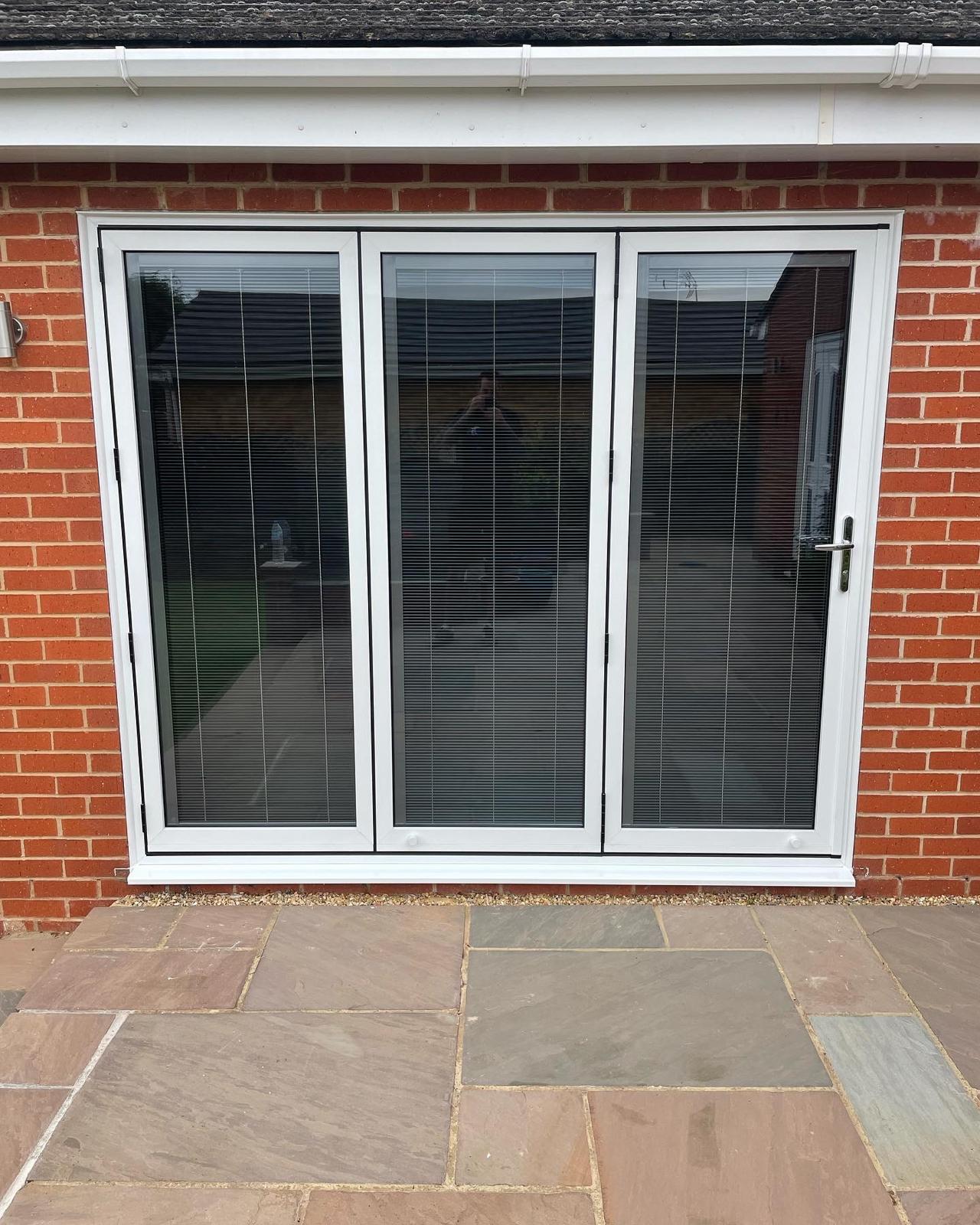 White UPVC three leaf bi-fold doors with integral blinds in a red brick bungalow wall opening onto a sandstone patio in Peterborough